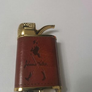 May include: A brown leather lighter with a gold trim and a pipe-shaped spout. The lighter has a John Walker logo embossed on the front.