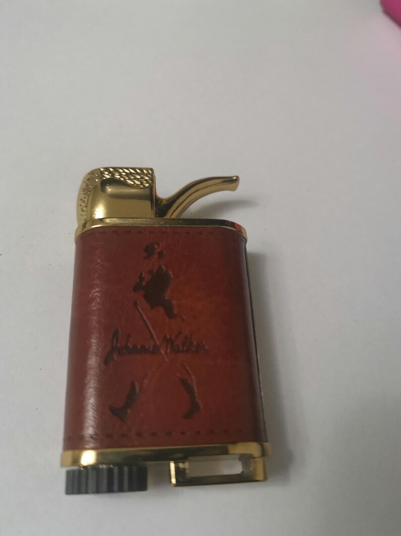 May include: A brown leather lighter with a gold trim and a pipe-shaped spout. The lighter has a John Walker logo embossed on the front.