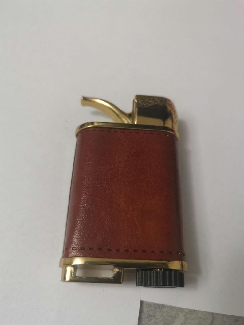 May include: A brown leather lighter with a gold trim and a gold-colored spout. The lighter has a black wheel on the bottom.
