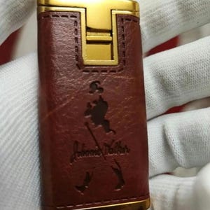 May include: A brown leather lighter with a gold trim and the John Walker & Sons logo embossed on the front.