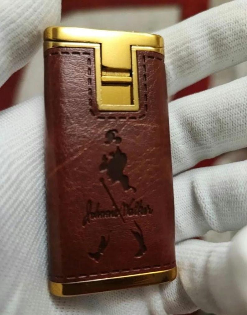 May include: A brown leather lighter with a gold trim and the John Walker & Sons logo embossed on the front.