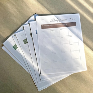 Puede incluir: Una pila de hojas de papel blancas con el texto "Filipenses 2:14" y "Hacedlo todo sin murmuraciones ni discusiones." Cada hoja tiene secciones para un nombre y fechas. Las hojas están sobre una superficie marrón claro.