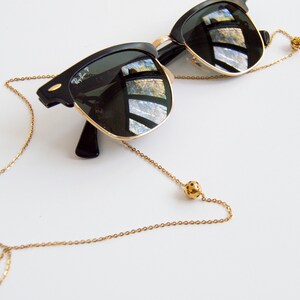 May include: A gold chain with two small gold balls on the ends. The chain is attached to a pair of black sunglasses.
