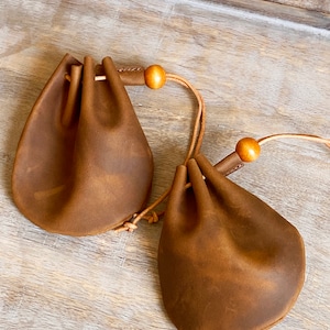 May include: Two brown leather drawstring pouches with wooden beads on the cords. The pouches are empty and lying on a wooden surface.