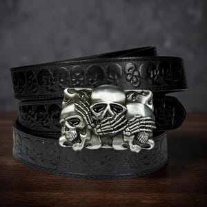 May include: A black leather belt with a silver skull buckle. The belt features embossed skulls along its length. The buckle has three skulls, with one covering its mouth with skeletal hands. A stylish accessory.