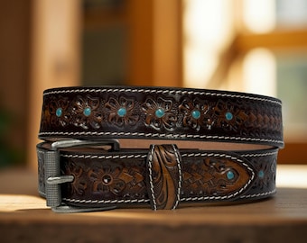 May include: A dark brown leather belt with floral and geometric embossed designs. The belt features turquoise-colored stone accents and a silver-colored buckle. White stitching outlines the edges of the belt.