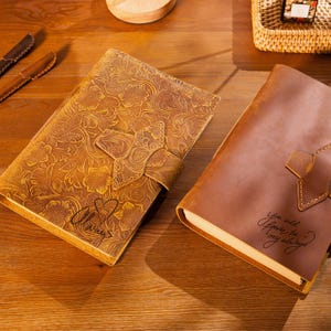 May include: Two leather journals with floral embossed designs. One journal is tan with a clasp, and the other is brown with a strap closure. The tan journal has the word "Always" inscribed, and the brown journal has the words "You will forever be my always."