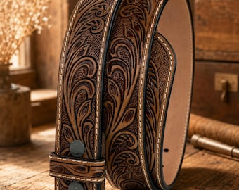 Puede incluir: Un cinturón de cuero marrón con intrincados grabados florales y patrones en relieve en toda la superficie. El cinturón incluye costuras blancas en contraste a lo largo de los bordes y dos broches metálicos oscuros en la trabilla frontal.