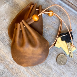 May include: Two brown leather drawstring pouches with wooden beads and a leather cord. A key and a coin are next to the pouches. The pouches have a rustic, worn appearance.