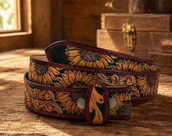May include: A leather belt featuring a detailed embossed sunflower pattern with yellow petals and dark centers against a black background. The belt has a reddish-brown edge finish and a small leather loop with two silver-toned metal rivets.
