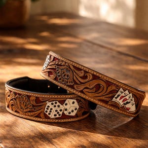 Op de afbeelding: Twee bruine leren hondenhalsbanden met gedetailleerde bloemen- en gokmotieven. Eén halsband toont dobbelstenen, de andere speelkaarten. De halsbanden hebben een donkerrode binnenkant en liggen op een houten oppervlak.