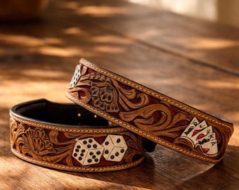 May include: Two brown leather dog collars with intricate floral and gambling-themed designs. One collar features dice, while the other displays playing cards. The collars have a dark red interior and are set against a wooden surface.