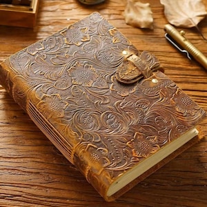 May include: A brown leather journal with an embossed floral design. The journal has a strap closure and gold-colored accents. The pages are visible, and the journal is on a wooden surface.