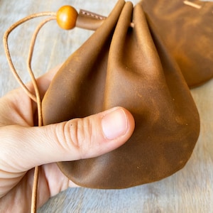 May include: A brown leather drawstring pouch with a wooden bead on the cord. The pouch is made of a soft, supple leather and has a rustic, vintage look.