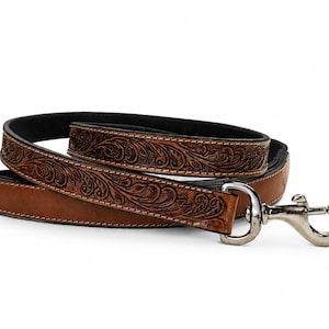 May include: A coiled brown leather dog leash featuring an intricate floral scrollwork pattern embossed along the strap. The leash includes a polished silver-toned metal swivel snap hook attached to one end, with black edges and visible stitching.