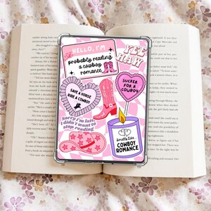 Puede incluir: Una funda transparente para teléfono con un fondo a cuadros rosa y blanco, adornada con varias pegatinas rosas. Las pegatinas presentan botas de vaquero, corazones y frases como "Hello, I'm probably reading a cowboy romance."