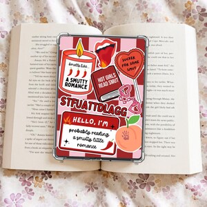 Könnte beinhalten: Eine transparente Handyhülle mit verschiedenen Aufklebern, darunter eine Kerze, Lippen, ein Buch und ein Pfirsich. Die Aufkleber tragen Sätze wie "A Smutty Romance" und "Hot Girls Read Smut". Die Hülle liegt auf einem Buch.