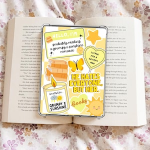Puede incluir: Un libro abierto con una funda transparente para teléfono con pegatinas amarillas. Las pegatinas incluyen texto que dice "Hello, I'm probably reading a grumpy x sunshine romance", una pila de libros, una mariposa y una vela.