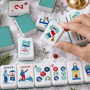May include: A set of rectangular mahjong tiles with white bases and teal backs, featuring winter-themed illustrations including skiers, a lift ticket, a scarf, mittens, and a lantern. Some tiles display the word JOKER and various numbers.