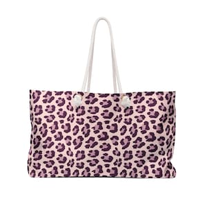 Peut inclure: Un grand sac fourre-tout avec un motif léopard rose. Le sac a des poignées en corde blanche et des accents blancs. L'imprimé léopard présente des taches violet foncé sur un fond rose clair. Le sac est conçu pour transporter des articles.