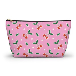 Könnte beinhalten: Rosa Reißverschlusstasche mit einem sich wiederholenden Muster aus roten Kirschen, grünen Blättern und weißen Sternformen. Die Tasche hat einen schwarzen Reißverschluss und einen goldfarbenen Zipper.