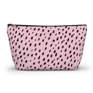 Könnte beinhalten: Eine rosa Kosmetiktasche mit schwarzem Reißverschluss und goldenem Zug. Die Tasche ist mit einem Muster aus schwarzen und magentafarbenen Pinselstrichen bedeckt. Die Tasche hat eine Trapezform.