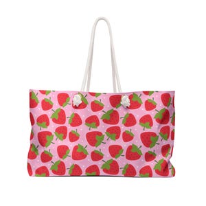 May include: A large tote bag with a repeating pattern of red strawberries with green leaves on a light pink background. The bag has white rope handles and white accents.