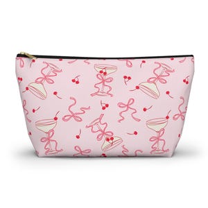 Può includere: Pochette cosmetica rosa con cerniera nera, con un motivo di calici di champagne, ciliegie e fiocchi. I calici sono decorati con ciliegie e legati con fiocchi rosa.
