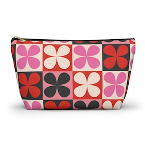 Può includere: Un beauty case con cerniera e motivo floreale retrò. Il design presenta una griglia di quadrati alternati in rosso, nero, rosa e crema, ciascuno con un fiore a quattro petali. La cerniera è color oro.