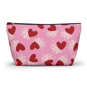 May include: A pink zipped pouch featuring a pattern of red hearts and white scribbled flower shapes. The pouch has a black zip and a gold-coloured zip pull.