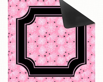 May include: A square mat with a pink background featuring a repeating pattern of white spiderwebs and small black spiders. A thick black border frames the central area, and the top right corner is folded over to reveal a solid black underside.