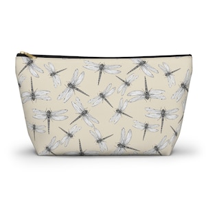 May include: Beige zippered pouch with a black and white dragonfly pattern. The pouch has a black zipper and a gold-colored zipper pull. The dragonfly design is repeated across the surface of the pouch.
