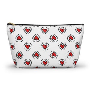 Può includere: Pochette per cosmetici bianca con un motivo ripetuto di cuori rossi con contorni a puntini neri. La borsa ha una cerniera dorata e un tirazip nero. È una pochette per cosmetici.