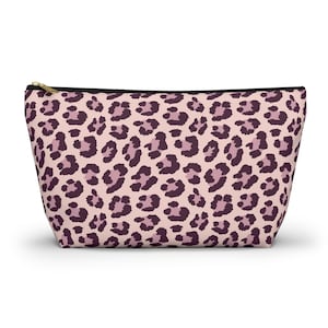 Peut inclure: Une trousse de maquillage rose avec un motif léopard dans les tons violets. Le sac a une fermeture éclair noire et une tirette dorée.