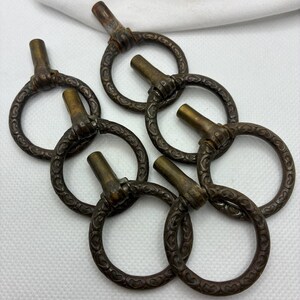 May include: Close-up of six antique bronze-colored ring pulls. Each ring has a decorative pattern and a cylindrical stem. The pulls are arranged in a cluster, suggesting they are for furniture or decorative use.