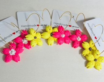 May include: Four pairs of handmade earrings featuring pink and yellow flower charms with gold hoops. The earrings are labeled "Handmade with love".