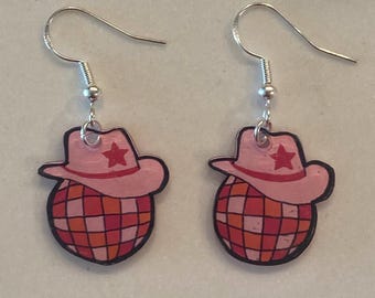 May include: A pair of silver hoop earrings with pink and red disco ball charms topped with pink cowboy hats with red stars.