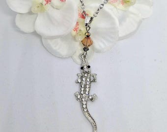 May include: A silver-toned metal necklace featuring a pendant shaped like a lizard or alligator, fully encrusted with clear sparkling rhinestones and two small black eyes. The chain includes a single faceted amber-colored crystal bead accent.