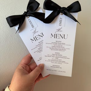 May include: Two vertical white paper menus with black text and a black satin ribbon bow at the top. Each menu features the names Brodey and Emily on a small tag attached to the ribbon. The text lists mains, sides, salad, and dessert options.