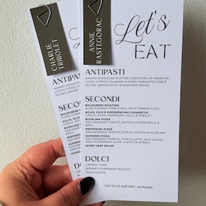 May include: Two white rectangular menu cards with black text titled Let's Eat, featuring sections for Antipasti, Secondi, and Dolci. Each card has a dark grey rectangular name tag attached with a silver paperclip, reading Charlie Tribolet and Annie Rastegorac.