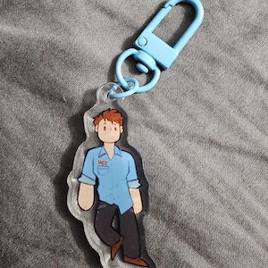 Op de afbeelding: Een heldere acryl sleutelhanger met een cartoon illustratie van een persoon in een blauw overhemd en donkere broek. De sleutelhanger heeft een lichtblauwe karabijnhaak en ring. De persoon heeft bruin haar en draagt een shirt met de tekst "WG2".