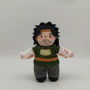 Puede incluir: Un muñeco de peluche que representa a un personaje con cabello negro, una cinta marrón y barba. El muñeco viste una túnica verde con un cinturón color canela, pantalones gris oscuro y vendas blancas en los brazos con detalles en color naranja.