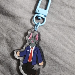 Könnte beinhalten: Ein klarer Acryl-Schlüsselanhänger mit einer cartoonartigen fledermausartigen Figur in einem blauen Blazer, einer roten Krawatte und einer schwarzen Hose. Der Schlüsselanhänger hat einen hellblauen Verschluss und Ring. Die Figur hat ein graues Gesicht und rosa Ohren.