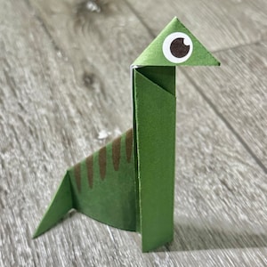 Può includere: Un dinosauro di origami verde con macchie marroni e un occhio bianco con una pupilla nera.