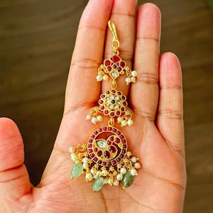 May include: A gold-toned headpiece with red and white gemstones, small pearls, and green accents. The design features a floral motif and a circular pendant with a peacock-like design. The headpiece is likely for special occasions.