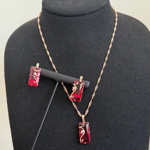 May include: A jewelry set featuring a necklace and earrings. The set includes a gold-colored chain necklace with a rectangular pendant and matching earrings. The pendant and earrings have a red stone with a decorative gold-colored design.