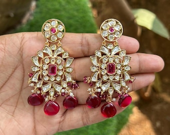 May include: A pair of gold earrings with red and white stones. The earrings have a square design with a large red stone in the center. The earrings are hanging from a small round piece with a smaller red stone in the center.