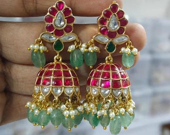 May include: A pair of gold-toned earrings with a floral design at the top, featuring red and clear gemstones. The earrings have a bell-shaped bottom with red and clear accents, and green and white bead dangles.
