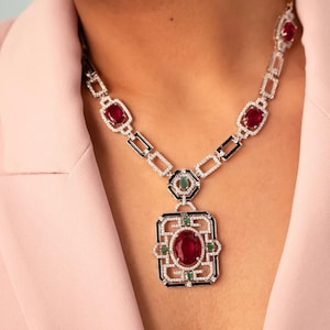 May include: A silver necklace with a large, oval, red gemstone pendant. The necklace features smaller red gemstones, emeralds, and black and white accents. The necklace is worn on a person wearing a pink blazer.