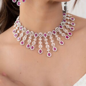 May include: A delicate silver necklace with a cascading design of ruby and diamond accents. The necklace features a central row of ruby stones with smaller diamonds surrounding them. The ruby stones are pear-shaped and the diamonds are round.
